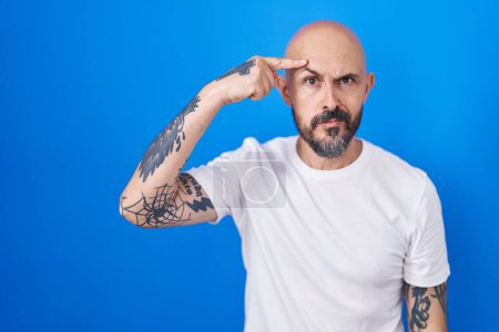 Foto de Hombre hispano con tatuajes de pie sobre fondo azul apuntando infeliz a la espinilla en la frente, fea infección de la cabeza negra. acné y problemas de piel - Imagen libre de derechos