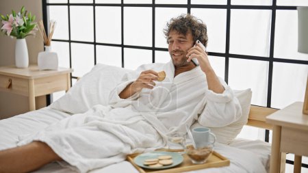 Foto de Hombre hispano joven desayunando hablando en smartphone en el dormitorio - Imagen libre de derechos