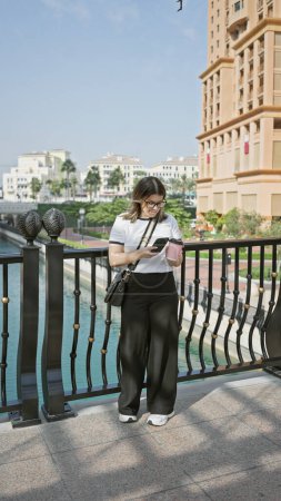 Téléchargez les photos : Une jeune femme vérifie son téléphone à doha, mettant en valeur l'architecture moderne et l'horizon arabe tranquille. - en image libre de droit