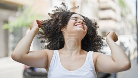 Foto de Joven hermosa mujer hispana sonriendo con confianza peinando el pelo con las manos en la calle - Imagen libre de derechos