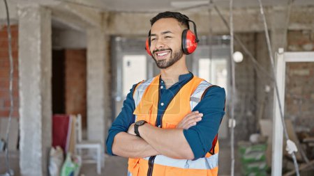 Photo for Young hispanic man builder with crossed arms wearing noise cancelling earmuffs at construction site - Royalty Free Image