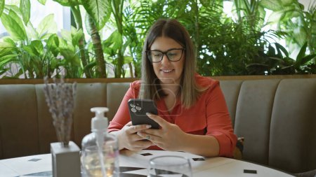 Téléchargez les photos : Une jeune femme brune en chemisier rouge assise dans un café à l'aide d'un smartphone - en image libre de droit