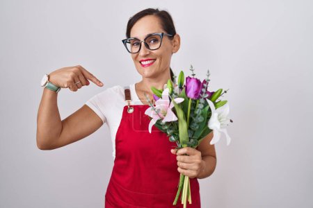 Téléchargez les photos : Femme brune du moyen âge portant un tablier travaillant dans un magasin de fleuristes tenant un bouquet avec un sourire sur le visage, se montrant du doigt fier et heureux. - en image libre de droit