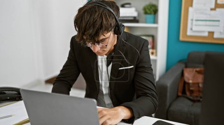 Photo for Young, handsome hispanic teenager hustling at the office, rocking headphones, engaged in professional business work, laser-focused on laptop success. - Royalty Free Image