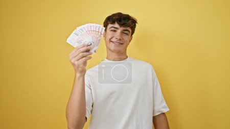 Ein fröhlicher junger hispanischer Teenager hält isländische Kronen-Banknoten in der Hand und strahlt Zuversicht aus, während er vor einem gelben, isolierten Hintergrund steht