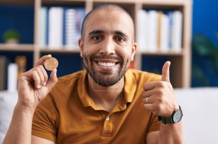 Téléchargez les photos : Homme hispanique avec barbe tenant monnaie virtuelle Bitcoin sourire heureux et positif, pouce vers le haut faire excellent et signe d'approbation - en image libre de droit