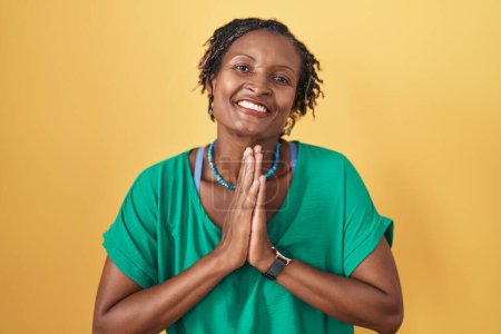 Téléchargez les photos : Femme africaine avec dreadlocks debout sur fond jaune priant avec les mains ensemble demandant pardon souriant confiant. - en image libre de droit