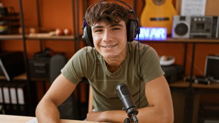 Téléchargez les photos : Jeune adolescent hispanique confiant, un musicien en herbe souriant, se délectant de son passe-temps alors qu'il porte des écouteurs dans un studio de radio professionnel, interprétant sa propre douce mélodie. - en image libre de droit