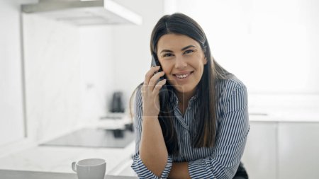 Foto de Joven mujer hispana hermosa hablando por teléfono apoyada en el mostrador de la cocina - Imagen libre de derechos