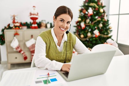 Photo for Young beautiful hispanic woman call center agent sitting by christmas tree working at home - Royalty Free Image