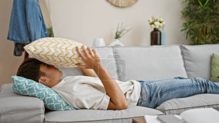 Photo for Pensive young hispanic teenager, lying indoors on a living room sofa, cushion on head, worried look in his home's interior - Royalty Free Image