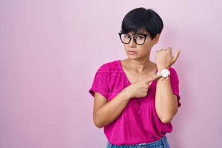 Téléchargez les photos : Jeune femme asiatique aux cheveux courts debout sur fond rose à la hâte pointant vers le temps de montre, impatience, regardant la caméra avec une expression détendue - en image libre de droit