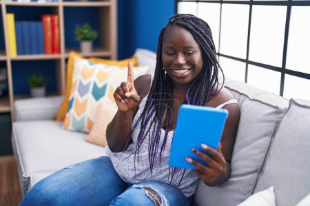 Foto de Mujer africana joven usando touchpad sentado en el sofá sonriendo con una idea o pregunta señalando el dedo con la cara feliz, número uno - Imagen libre de derechos
