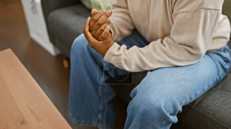 Foto de Mujer afroamericana agarrándose de las manos mientras está sentada en el sofá en el interior para un look de retrato relajado. - Imagen libre de derechos