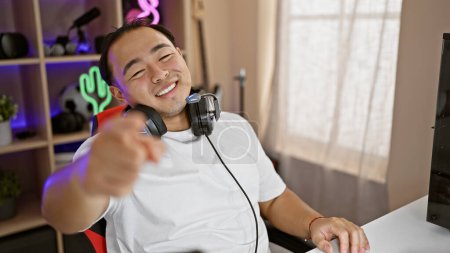 Téléchargez les photos : Beau jeune chinois, un joueur et streamer numérique vivant, pointant vers la caméra dans une salle de jeux sombre, au milieu du frisson du jeu nocturne, assis avec une console, casque sur - en image libre de droit