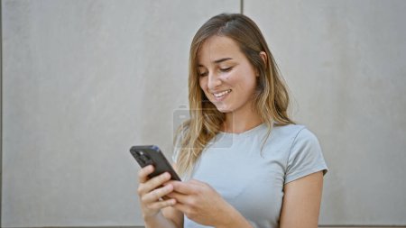Photo for Cheerful young blonde woman happily touching her smartphone, engaged in a digital conversation on a sunny city street - Royalty Free Image
