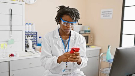 Femme scientifique afro-américaine en blouse de laboratoire en utilisant un smartphone en laboratoire