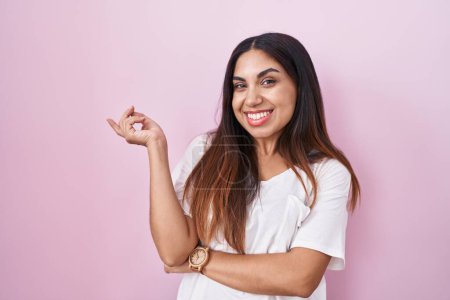 Téléchargez les photos : Jeune femme arabe debout sur fond rose avec un grand sourire sur le visage, pointant avec le doigt de la main vers le côté en regardant la caméra. - en image libre de droit