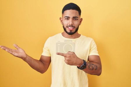 Foto de Young hispanic man standing over yellow background amazed and smiling to the camera while presenting with hand and pointing with finger. - Imagen libre de derechos