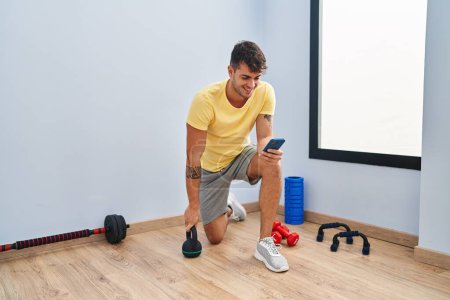 Téléchargez les photos : Jeune homme hispanique utilisant un smartphone en utilisant la formation kettlebell au centre sportif - en image libre de droit