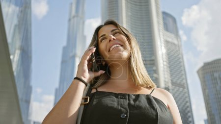 Photo for A smiling young woman talks on her phone in front of the towering skyscrapers of dubai under a blue sky. - Royalty Free Image