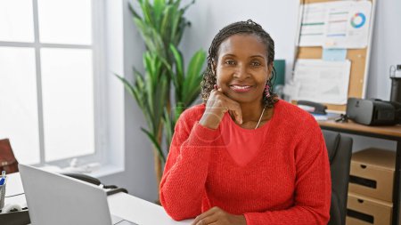 Foto de Mujer afroamericana sonriente vestida de rojo en una oficina brillante con computadora portátil, auriculares y planta. - Imagen libre de derechos