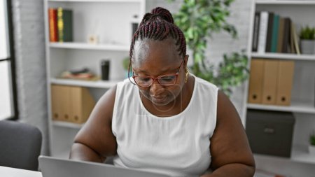 Téléchargez les photos : Travailleuse afro-américaine concentrée excellant dans les affaires. travaillant avec diligence à son ordinateur portable, cette dame patron incarne le succès et le professionnalisme dans son bureau. - en image libre de droit