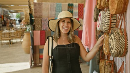 Téléchargez les photos : Femme souriante dans un chapeau de soleil explorer un marché traditionnel à dubai, en regardant des sacs artisanaux. - en image libre de droit
