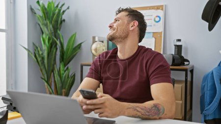 Foto de Hombre hispano guapo con barba sonriendo mientras usa el teléfono inteligente en la oficina moderna. - Imagen libre de derechos