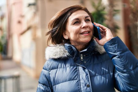 Téléchargez les photos : Femme d'âge moyen souriant confiant parler sur le smartphone à la rue - en image libre de droit