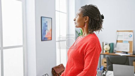 Téléchargez les photos : Femme afro-américaine dans un cadre de bureau regardant par la fenêtre, contemplant, portant des vêtements d'affaires décontractés. - en image libre de droit