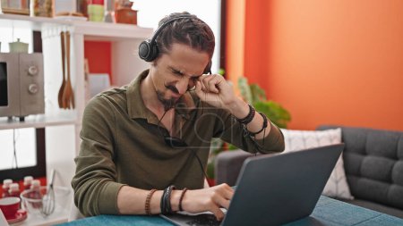 Téléchargez les photos : Jeune homme hispanique utilisant ordinateur portable et écouteurs fatigués à la salle à manger - en image libre de droit