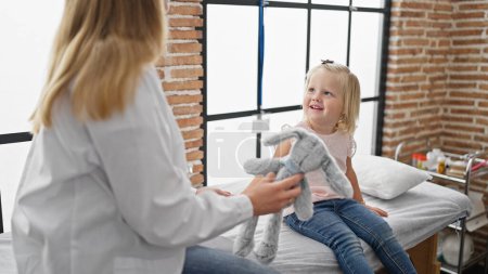Glückliche Kinderpatientin mit Hasenspielzeug in der Kinderklinik, fröhliche ärztliche Untersuchung bei der selbstbewussten Ärztin