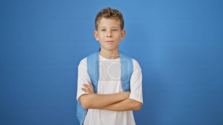 Photo for Adorable blond academic boy standing relaxed, arms crossed, showing serious concentration on his cute face against isolated blue wall background - Royalty Free Image