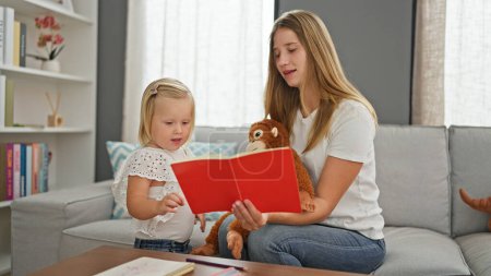 Photo for Serious caucasian mother and little daughter enjoy reading a book together, relaxing at home on the sofa surrounded by toys - Royalty Free Image