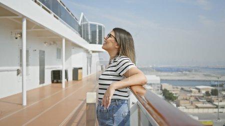 Foto de Una joven y sonriente mujer con gafas de sol y atuendo casual disfrutando de la vista al mar desde la cubierta de un crucero. - Imagen libre de derechos