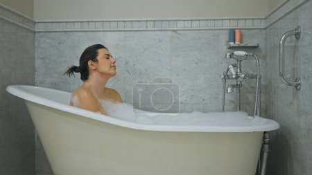 Photo for A relaxed woman enjoying a bubble bath in a clean, homely bathroom with bright lighting. - Royalty Free Image