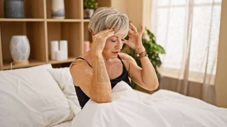 Photo for Mature woman with grey hair looking stressed in a bedroom setting, depicting health or mental well-being concepts. - Royalty Free Image