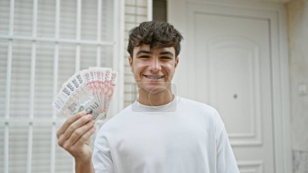 Cheerful young hispanic teenager confidently flashing a wad of iceland krona banknotes with a dazzling smile on a bustling city street, revealing an empowering expression of financial joy