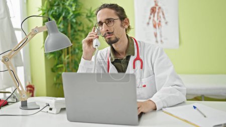 Téléchargez les photos : Jeune homme hispanique médecin utilisant un ordinateur portable parlant au téléphone à la clinique - en image libre de droit