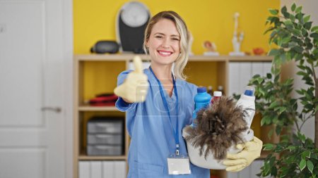 Téléchargez les photos : Jeune femme blonde nettoyant professionnel tenant des produits faisant geste pouce vers le haut au bureau - en image libre de droit