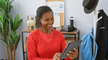 Téléchargez les photos : Une femme afro-américaine souriante dans un pull rouge utilisant une tablette dans un environnement de bureau avec des plantes et un porte-manteau. - en image libre de droit