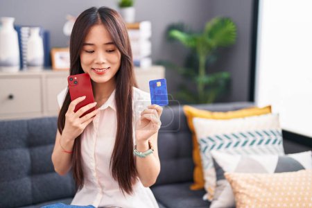 Téléchargez les photos : Jeune femme chinoise utilisant smartphone et carte de crédit assis sur le canapé à la maison - en image libre de droit