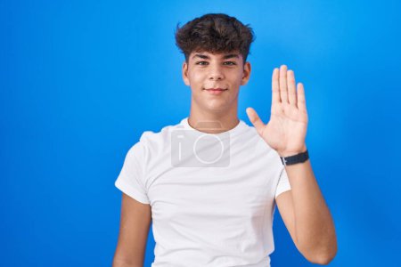 Téléchargez les photos : Adolescent hispanique debout sur fond bleu renonçant à dire bonjour heureux et souriant, geste de bienvenue amical - en image libre de droit