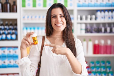 Téléchargez les photos : Jeune femme brune faisant du shopping à la pharmacie pharmacie tenant des pilules souriant heureux pointant avec la main et le doigt - en image libre de droit