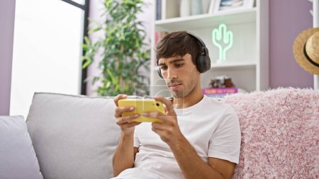 Photo for Young hispanic man playing video game sitting on sofa at home - Royalty Free Image