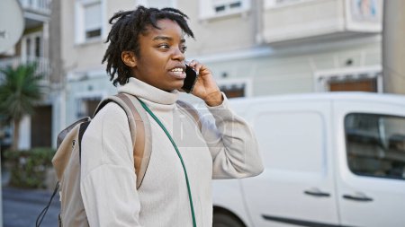 Photo for A young african american woman with dreadlocks talks on a phone while walking on an urban city street. - Royalty Free Image