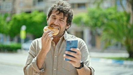 Téléchargez les photos : Jeune homme hispanique manger beignet en utilisant smartphone à la rue - en image libre de droit