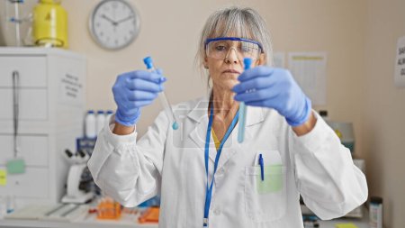 Une femme scientifique mature aux cheveux gris mène des expériences en laboratoire, tenant des éprouvettes avec du liquide bleu.
