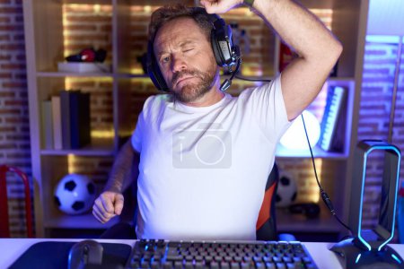 Téléchargez les photos : Homme d'âge moyen avec barbe jouant à des jeux vidéo portant des écouteurs étirant le dos, fatigué et détendu, endormi et bâillant pour tôt le matin - en image libre de droit